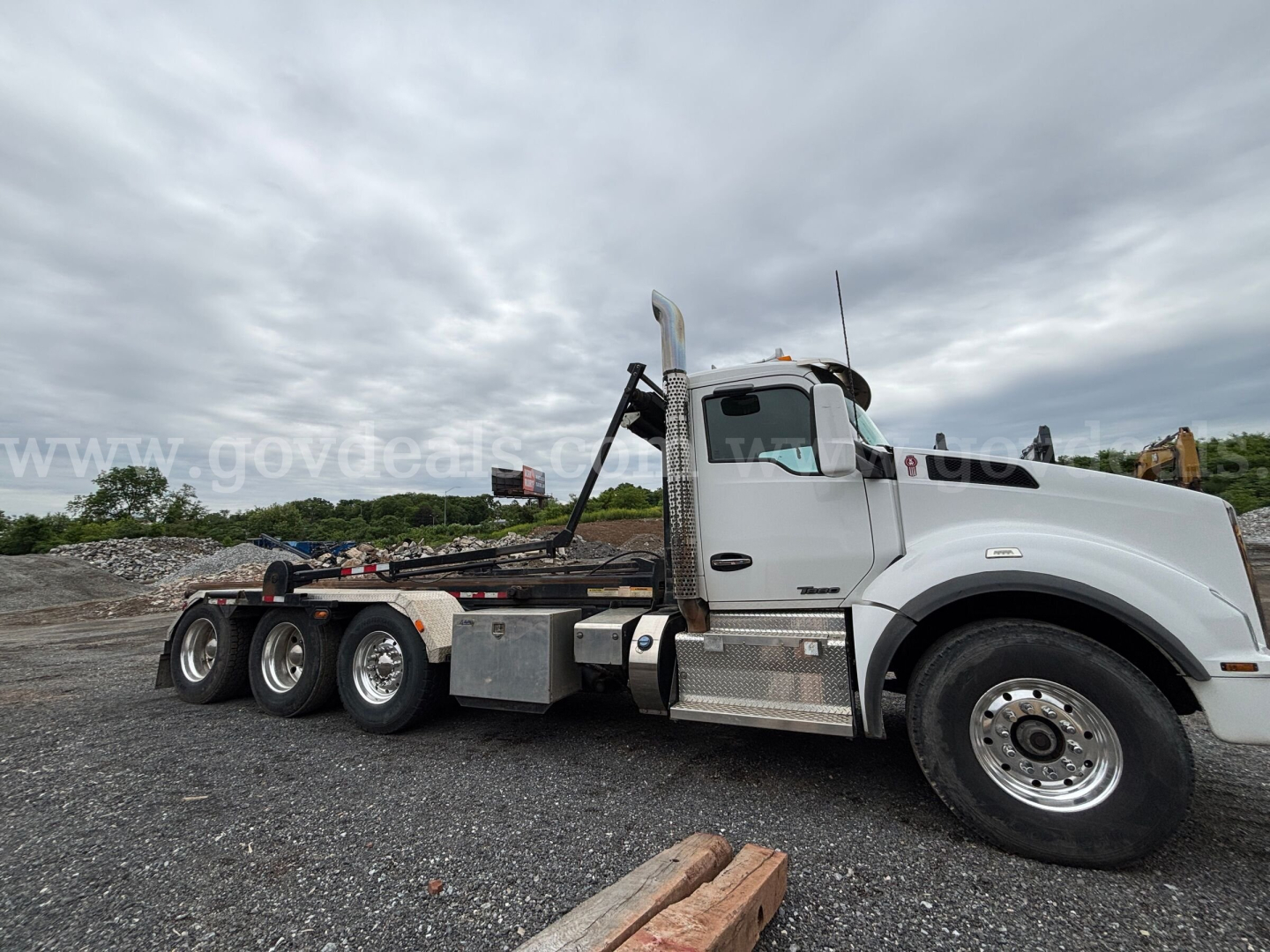 Kenworth T880 rolloff truck in side view showing cab, chrome fuel tanks, dual rear axles, and hydraulic hooklift system with metal toolbox and steps.
