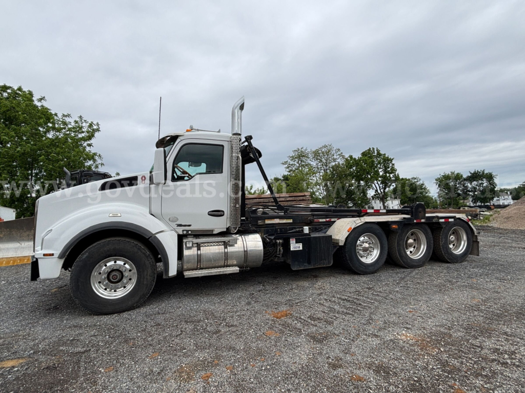 Kenworth T880 rolloff truck with white cab and long wheelbase, side view showing fuel tanks and exhaust stack.