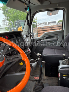 Kenworth T880 truck interior showing steering wheel, dashboard, gear shifter, and passenger seat; windshield with rearview mirror; hanging lanyard and cords; floor mat and control panels; label text not clearly legible.