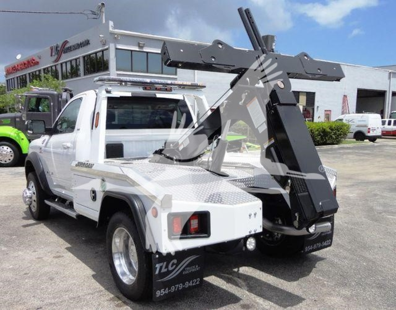Ram 4500 tow truck with large hydraulic boom and winch, white cab, mounted crane, rear view showing TLC contact info, dealership lot in background.