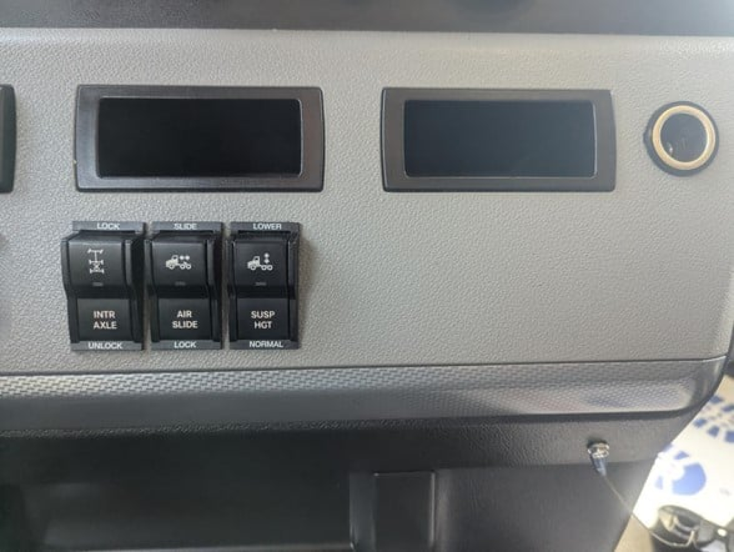 Dashboard panel showing rocker switches labeled LOCK INTR AXLE, A B R SLIDE, SUSP HLT, LOWER, etc. Two empty instrument recesses and a circular cigarette lighter/12V socket on Freightliner Cascadia dash.