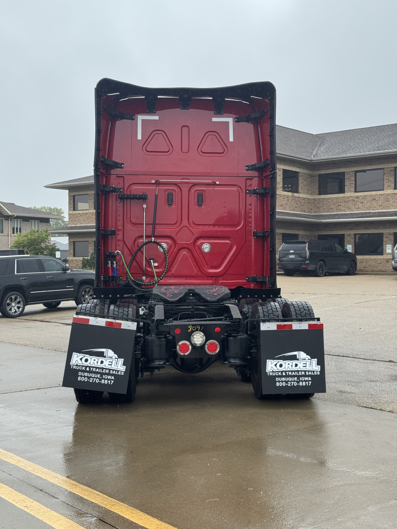 Rear view of a Freightliner Cascadia truck tractor, red cab interior visible through open trailer hitch area, black mud flaps with white Kirdele logo, concrete lot.