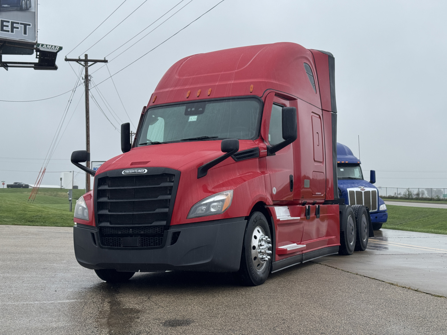 Red Freightliner Cascadia 126 sleeper cab tractor in a parking area, front-left angle, with chrome grille and black bumper, docking with blue trailer in background.