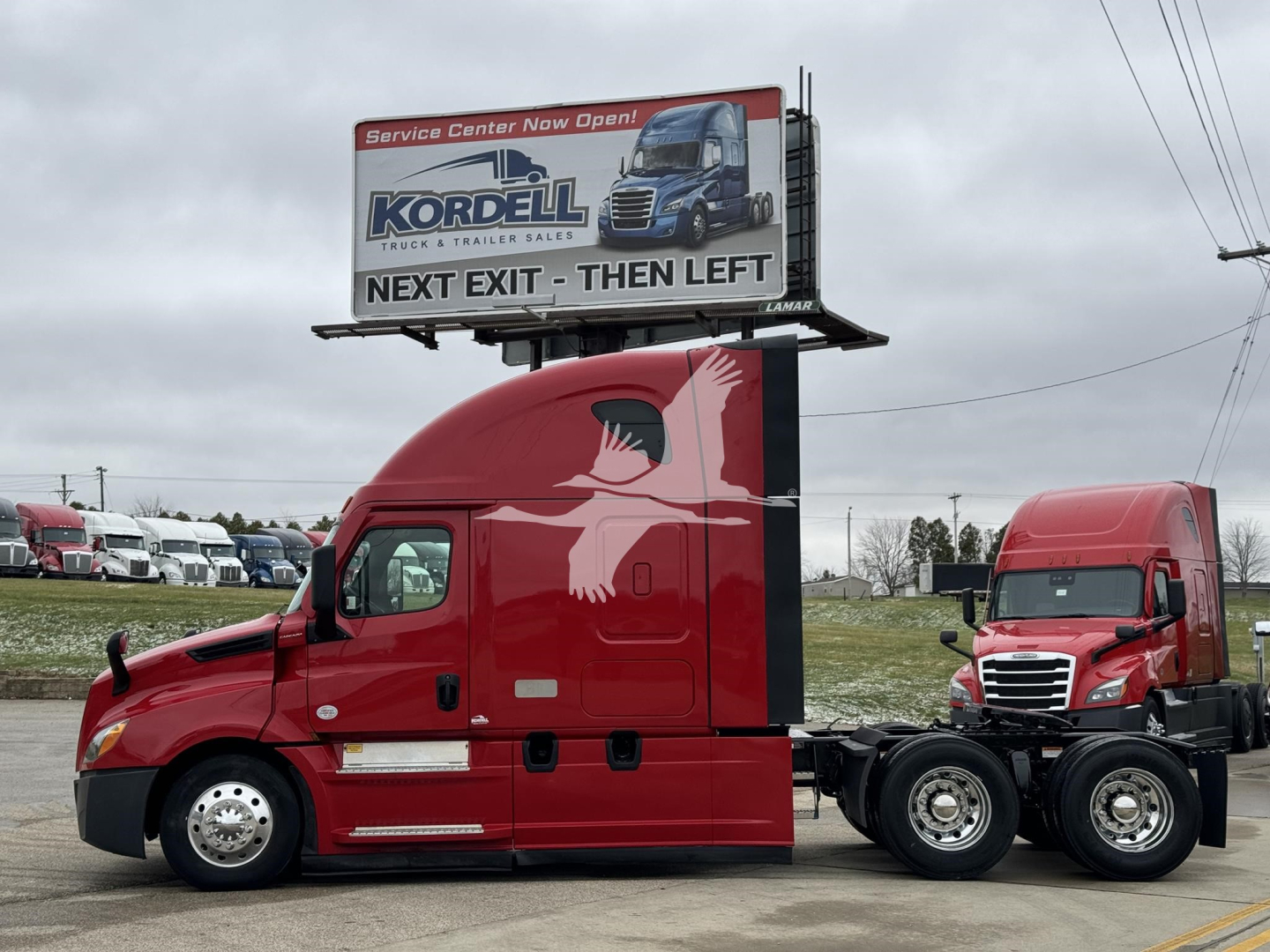 Freightliner Cascadia sleeper tractor unit in red, side view showing cab and chassis with sleeper, single hood, billboard behind it, other red trucks in background.