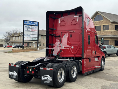 Truck rear-left view showing Freightliner Cascadia 126 sleeper cab with red exterior and dual rear axles; visible text on rear mudflaps includes Kordell Trucks logo and contact numbers; dealership sign in background.