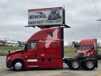 Freightliner Cascadia sleeper tractor unit in red, side view showing cab and chassis with sleeper, single hood, billboard behind it, other red trucks in background.