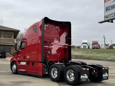 Truck cab: 2023 Freightliner Cascadia 126 with red exterior, sleeper cab, side view showing the rear chassis and dual rear axles.
