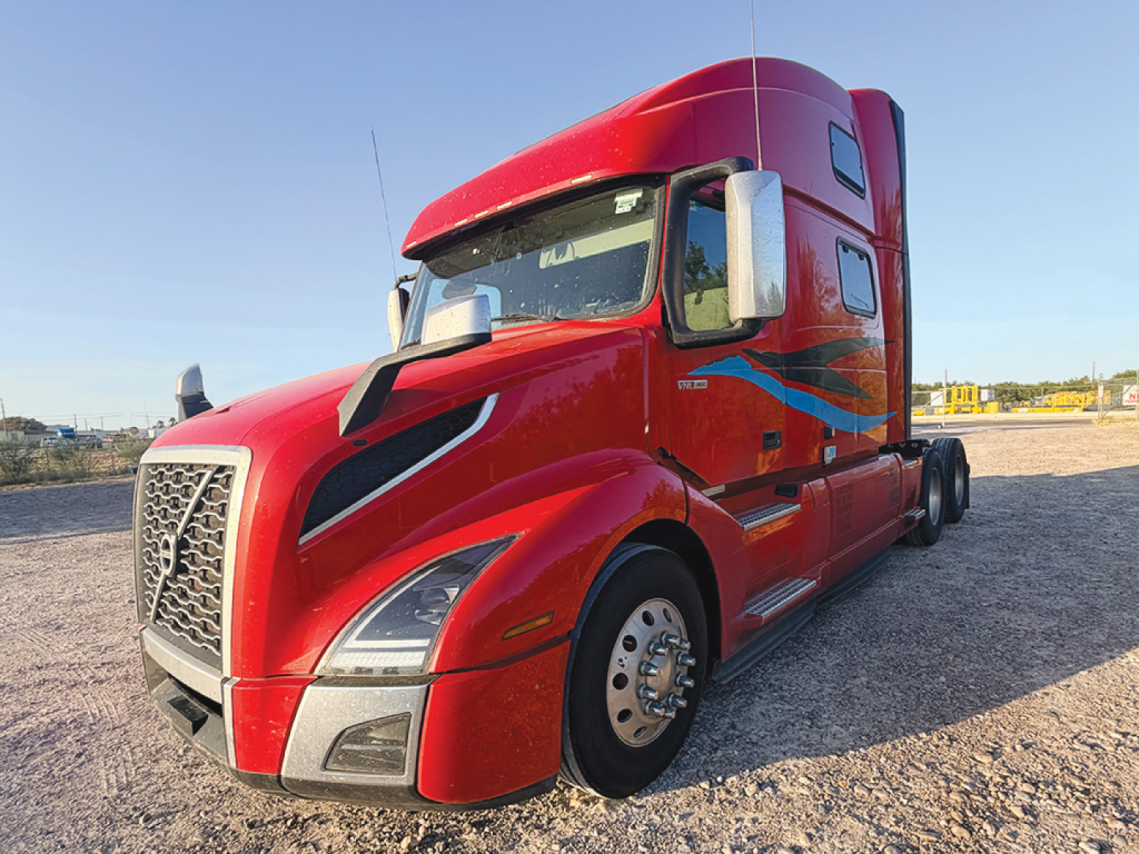 Truck cab in a bright red Volvo VNL 760 conventional sleeper, side view showing aerodynamic hood, large chrome grille, dual rear axles, and sleeper cab.