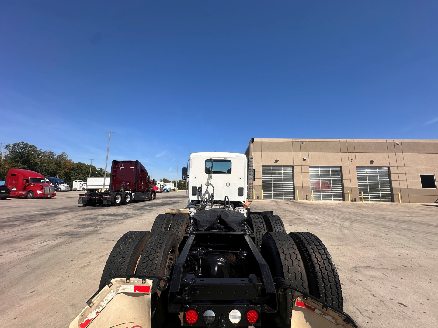 Kenworth T880 cab-over? Actually conventional daycab rear chassis with dual rear axles, showing fifth wheel and drivetrain. Visible text includes nothing readable on engines; truck yard with other trucks and building in background.