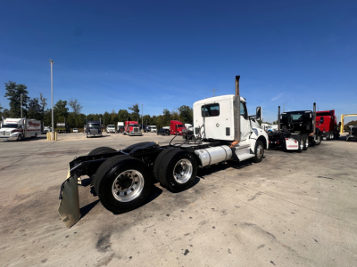 Kenworth T880 daycab tractor chassis with exposed rear axles and white cab, in a truck yard; various trucks in background.