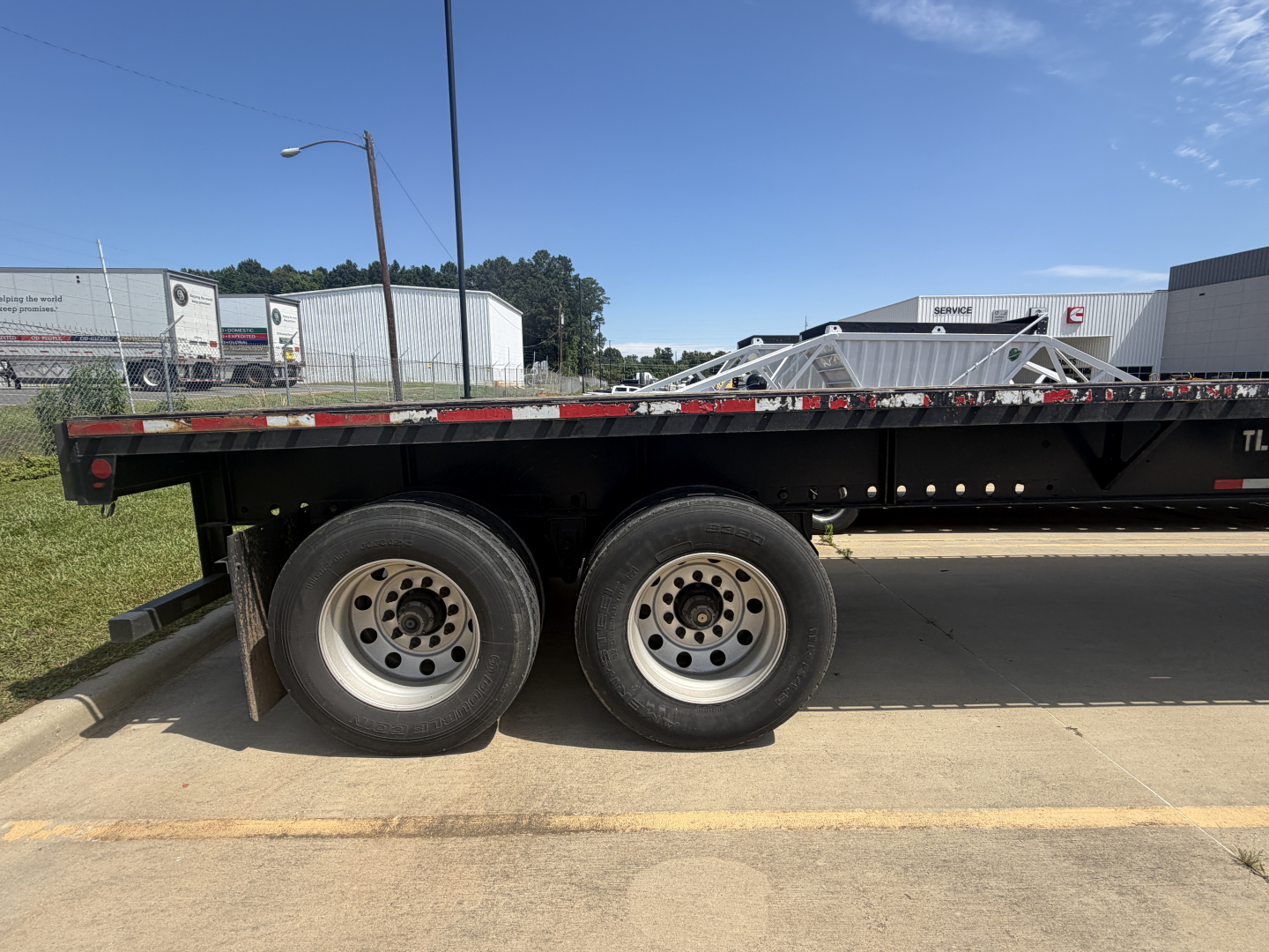 Flatbed trailer side with two wheels and spare; no visible text on the trailer side except small reflective tape. The image shows a Fontaine HAVSF12SST trailer with black deck and metal frame.