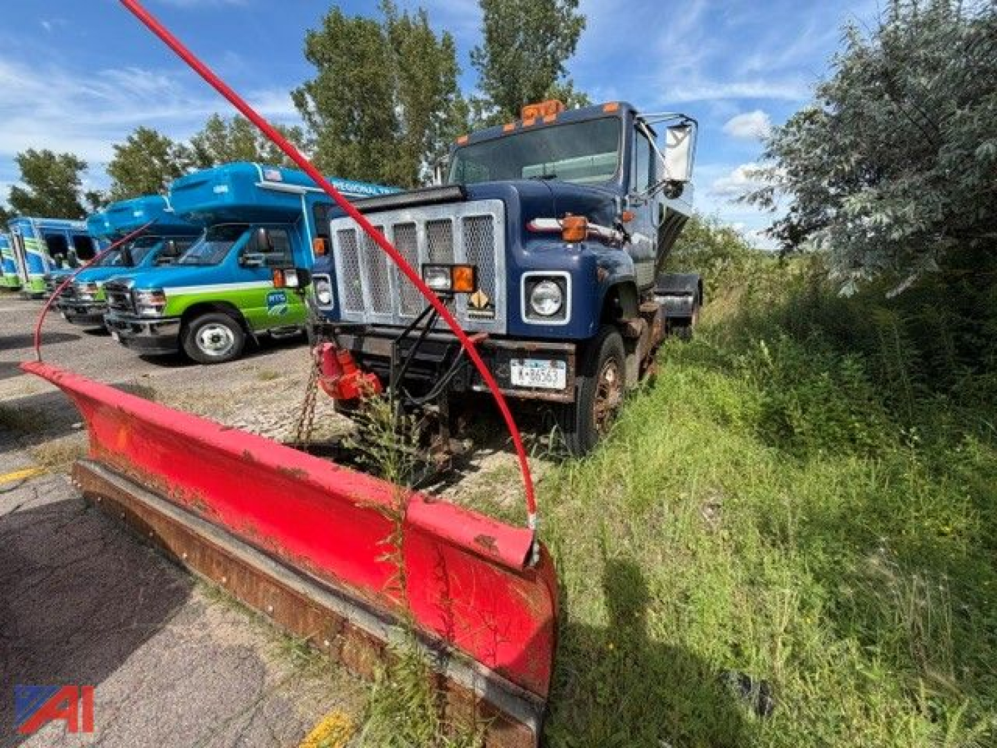 International 2574 truck with stone spreader and red blade in grassy lot; other equipment in background.
