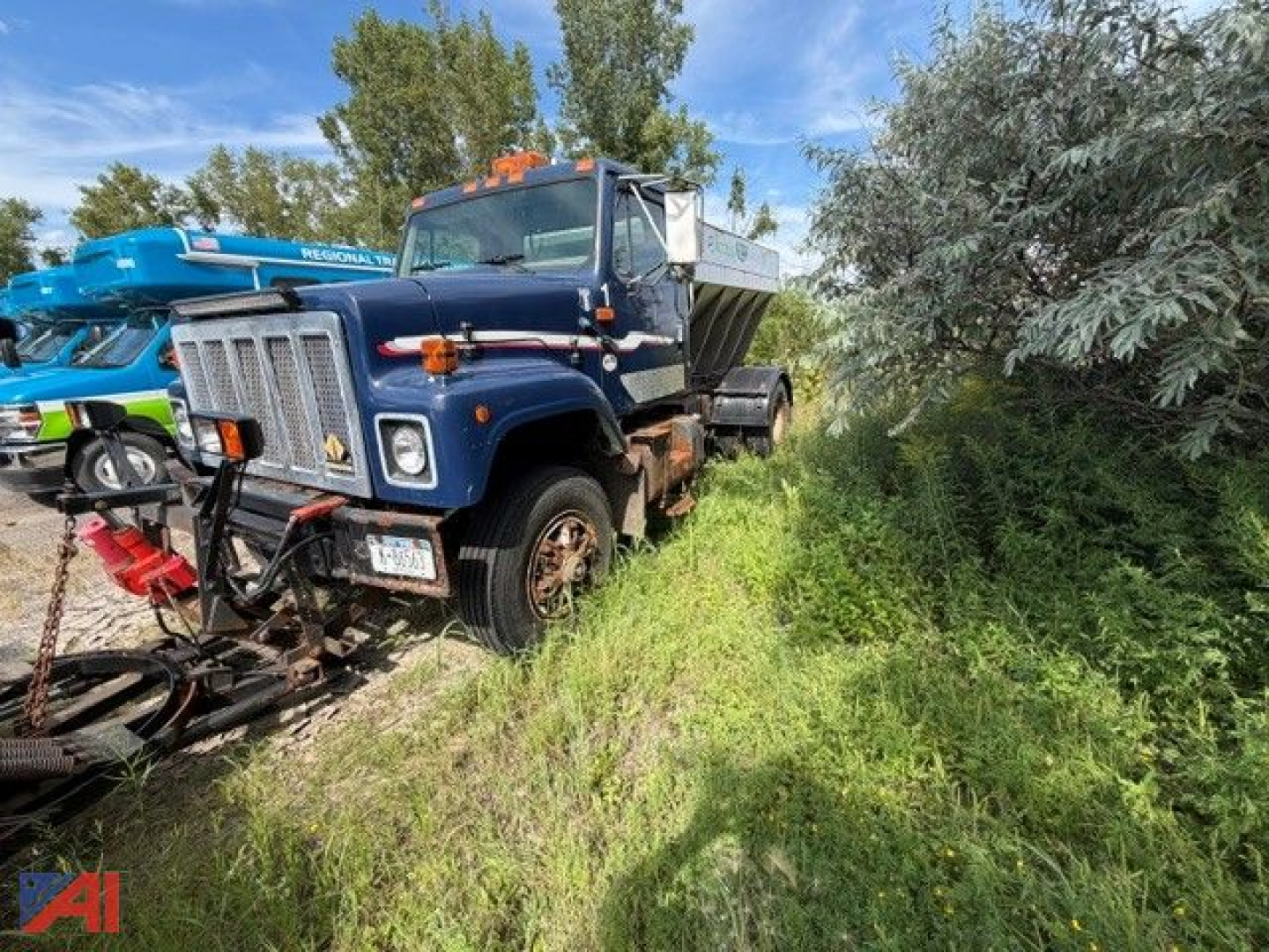Truck in a field showing a blue International 2574 stone spreader with a front grille, large side mirrors, and an orange beacon light; vegetation around and a red attachment in front.