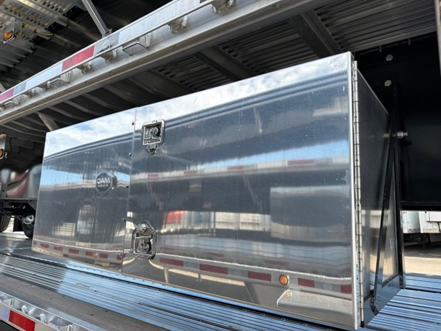 Flatbed trailer side storage box mounted under the flatbed, with polished aluminum diamond-plate door and latch hardware.