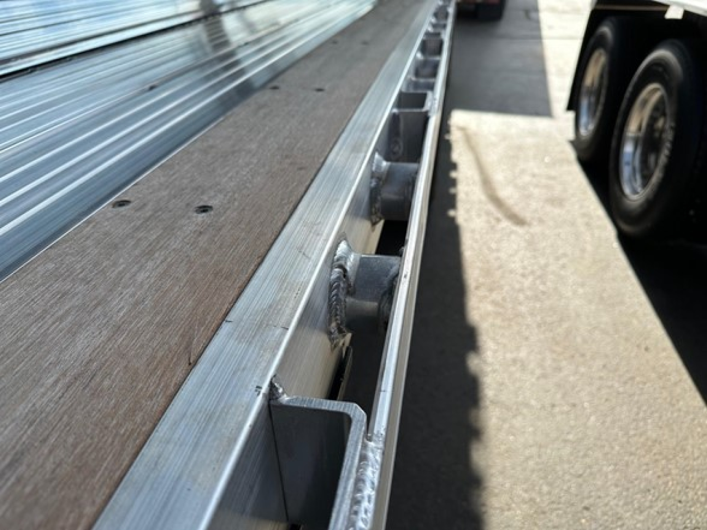 Trailer side rail and stake pocket with metal deck and riveted channels on a flatbed trailer; wheel well visible in background.