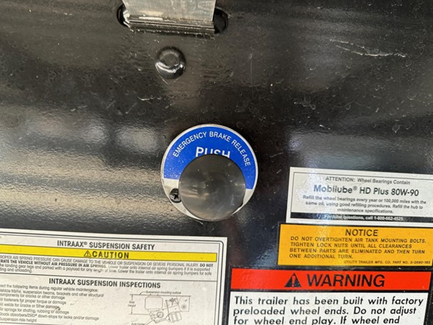 Trailer interior showing blue circular safety plug cover labeled Emergency Brake Release and various warning/inspection labels. Visible text includes INTRAX SUSPENSION SAFETY CAUTION and NOTICE, with a white label explaining wheel bearings