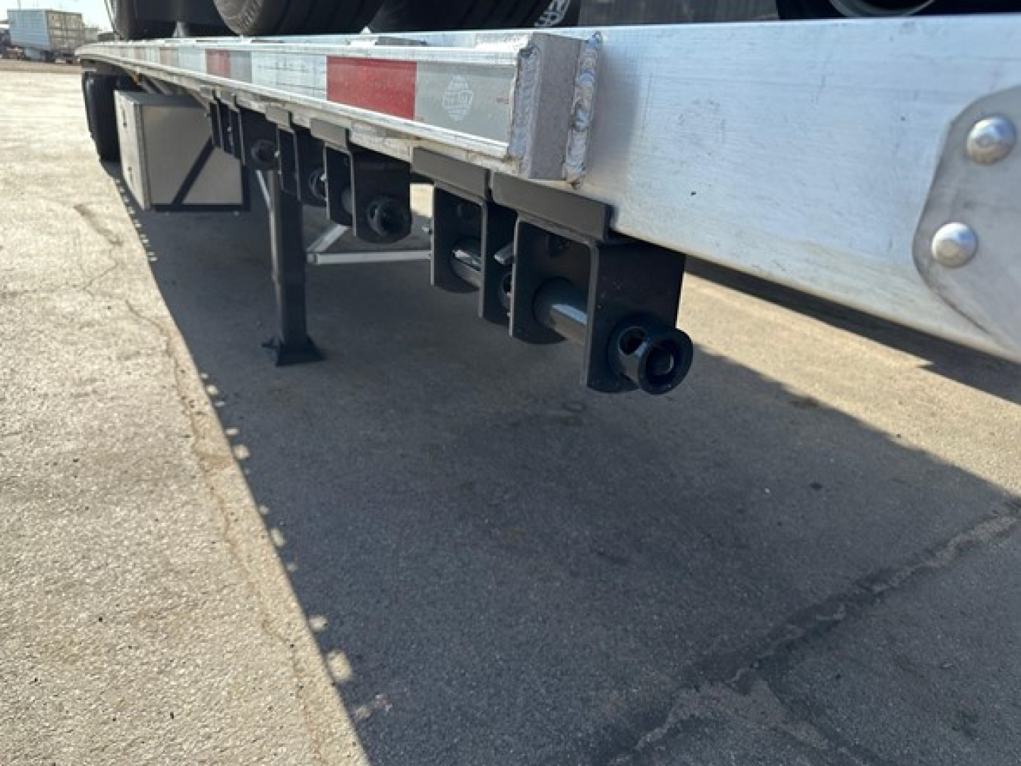 Utility flatbed trailer underside showing tools and attachment brackets, with black tubular hangers and support legs, silver frame, and rear underrun bar.