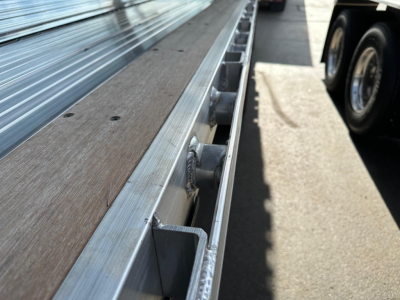 Flatbed trailer edge with metal side rails and wooden deck, wheel well visible in distance, showing structural brackets and riveted seams.