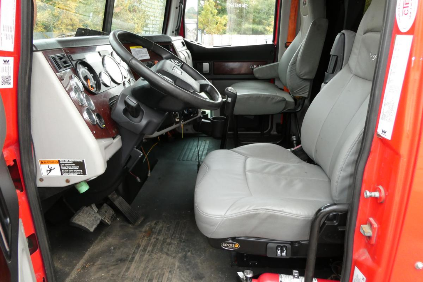 Truck cab interior of a 2019 Western Star 5700XE showing gray driver and passenger seats, steering wheel, dashboard, and pedals with worn floor mat.