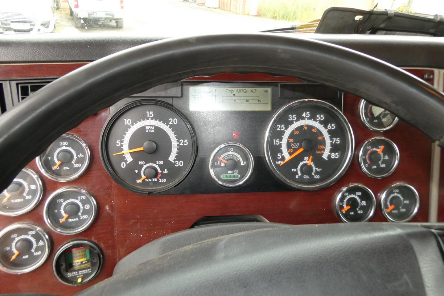 Truck cab dashboard with large analog gauges and steering wheel; several dials show speedometer, tachometer, fuel, air pressure, and other indicators; woodgrain panel around gauges.