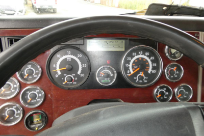 Truck cab dashboard with large analog gauges and steering wheel; several dials show speedometer, tachometer, fuel, air pressure, and other indicators; woodgrain panel around gauges.
