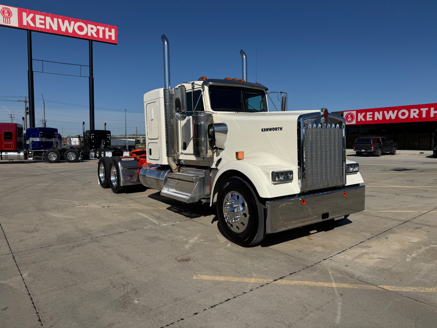 Kenworth W900L conventional sleeper truck in a dealership lot, white cab with chrome grille, dual tall exhaust stacks, chrome bumper, and Kenworth signs in the background.