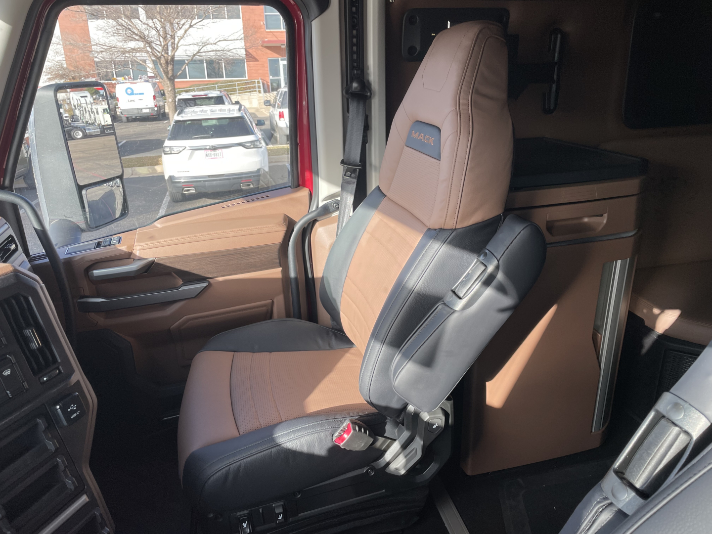 Truck cabin interior: driver's seat with Mack logo, brown and black seats, dashboard controls, side window, and sleeping area behind.