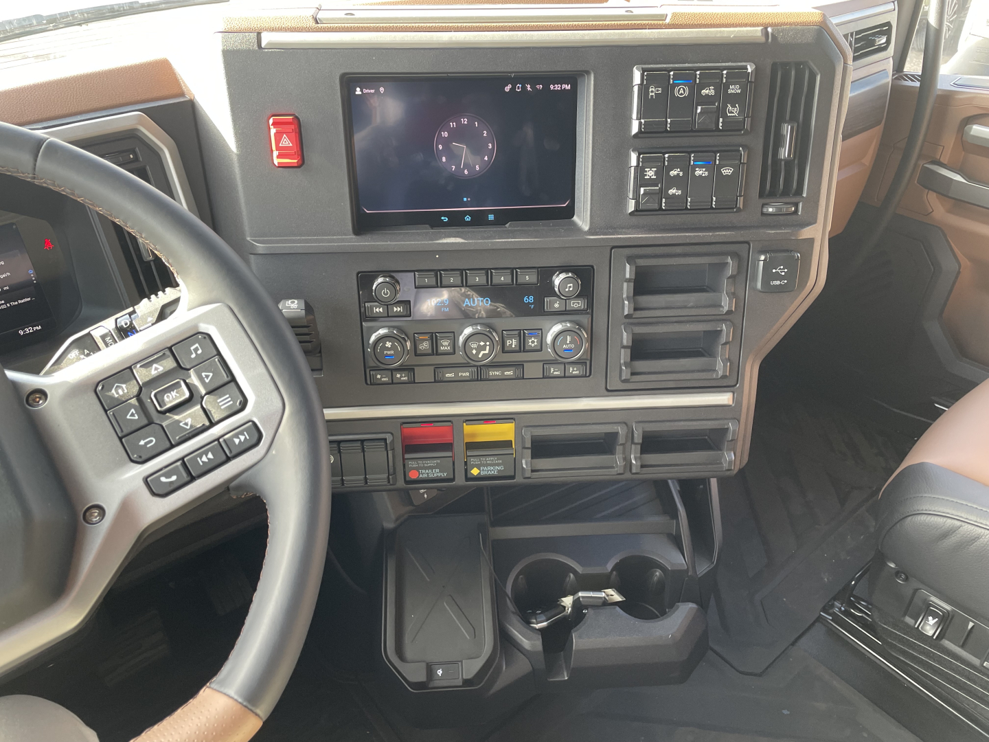 Dashboard and center console of a Mack PR64T76H truck showing steering wheel, touchscreen/instrument cluster, climate controls, switches, cup holders, and storage compartments.