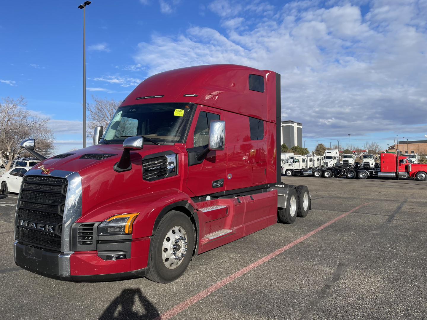 Mack PR64T76H sleeper cab semi truck in red, front-left side visible, with chrome grille and large, tall cab; background shows dealership lot with other trucks.