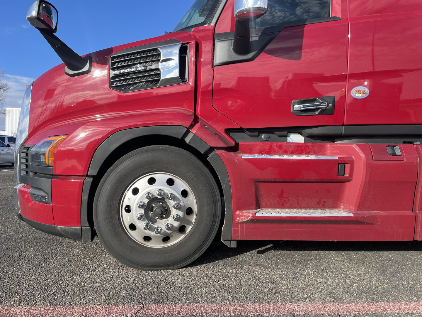 Mack PR64T76H truck side view showing front wheel, red cab, and sleeper, with chrome wheel hub and step.