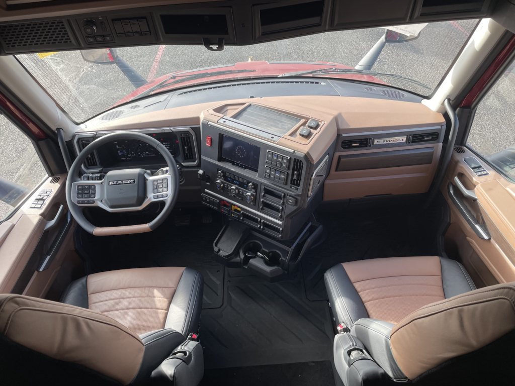 Cab interior of a new 2026 Mack PR64T76H truck showing tan and black seats, steering wheel, dashboard with control panels, gear shifter, and central console.