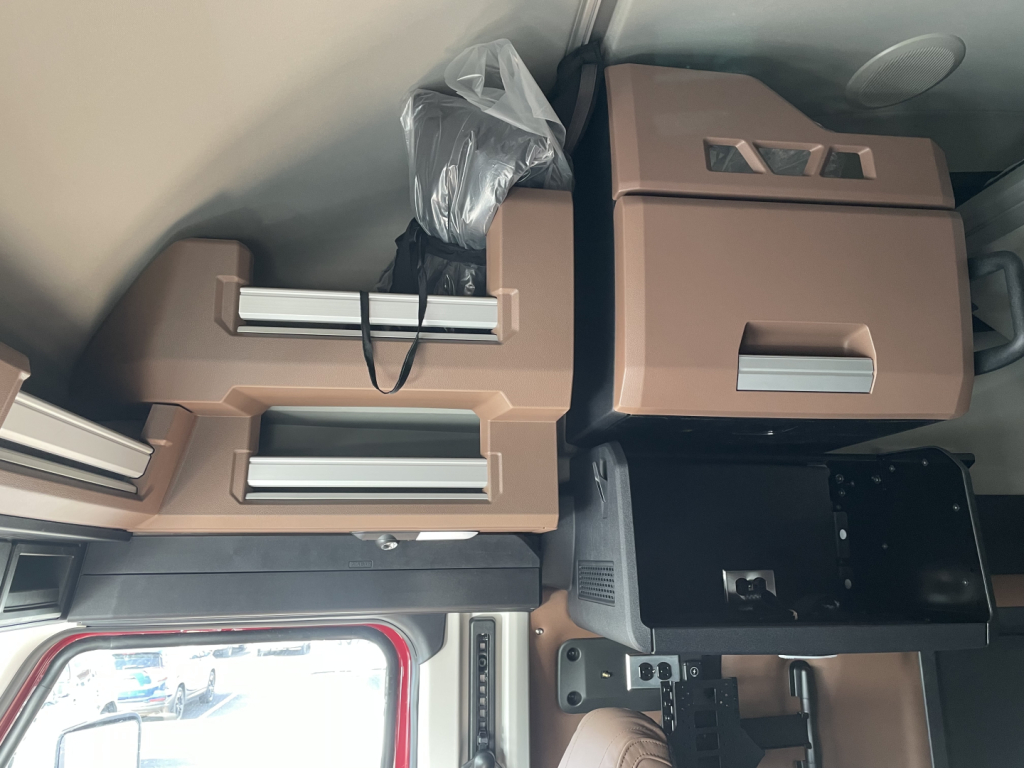 Cabin interior with tan storage compartments and a black control panel mounted on a wall, showing multiple drawer-like compartments and a plastic bag.