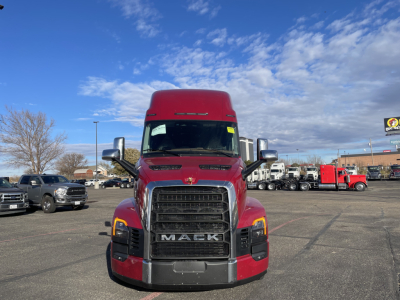 Mack semi truck front view in a lot showing bold black grille with MACK letters, red exterior, chrome bumper, large side mirrors, day cab-style with sleeper behind cab.