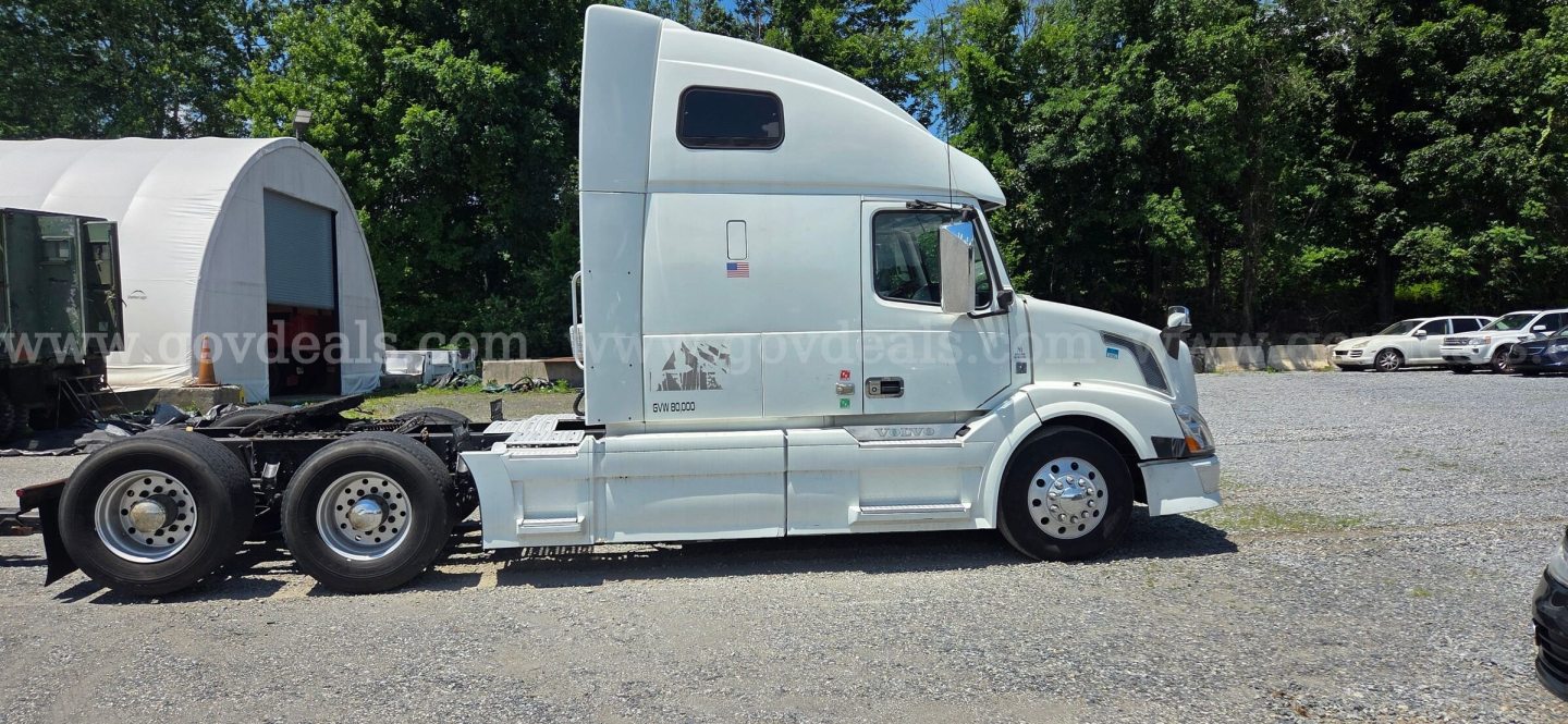 Semi tractor truck side view showing a light blue Volvo VNL with sleeper cab and attached trailer chassis. Visible text on cab includes model markings and side badges.