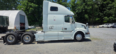 Semi tractor truck side view showing a light blue Volvo VNL with sleeper cab and attached trailer chassis. Visible text on cab includes model markings and side badges.