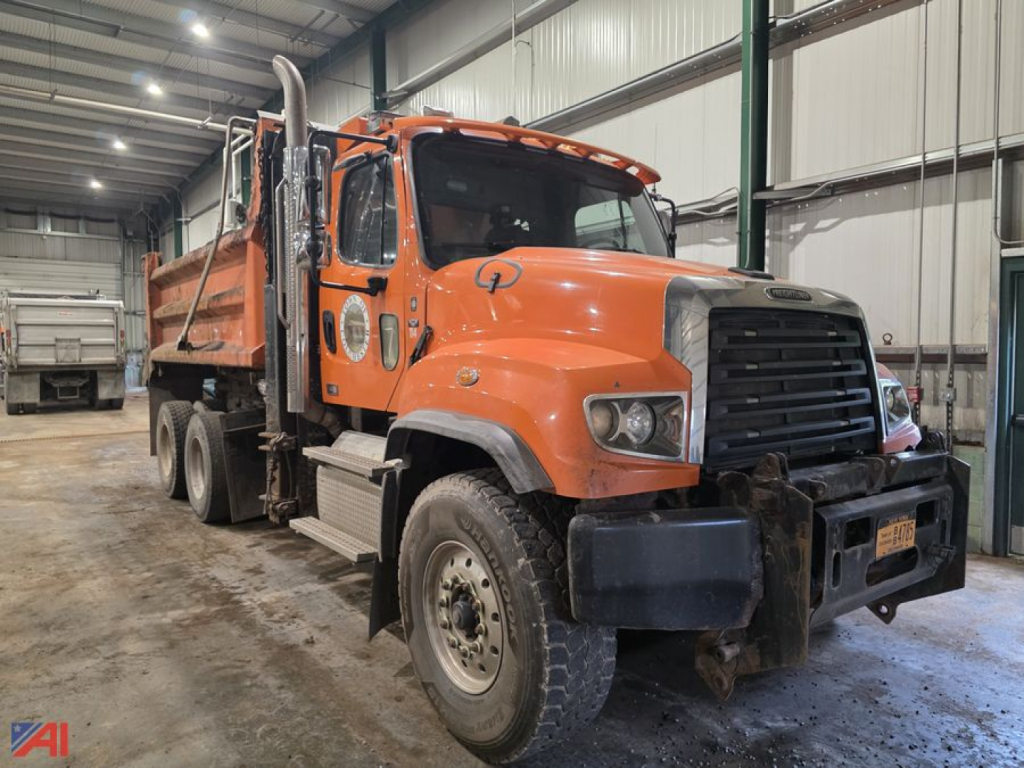 Dump truck cab and chassis in an indoor lot showing orange Freightliner 114SD with front grille, multiple axles, and dumping bed.
