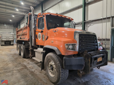 Dump truck cab and chassis in an indoor lot showing orange Freightliner 114SD with front grille, multiple axles, and dumping bed.