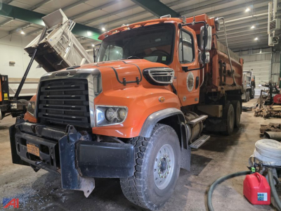 Orange Freightliner 114 SD dump truck in a workshop, front hood open, showing large grille, headlights, and front bumper with winch guard; dump bed raised slightly with interior bed visible.