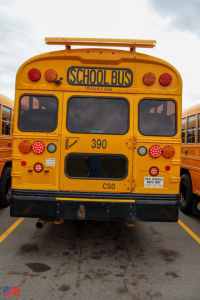 Back view of a yellow school bus with 'SCHOOL BUS' sign, number 390, CSD, and multiple red warning lights in a bus lot.