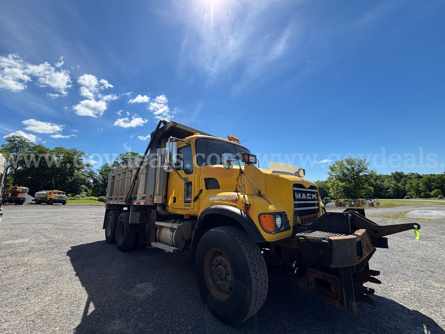 Mack CV713 dump truck in yard, front-right view, yellow cab with dump bed, large front bumper and snowplow attachment visible