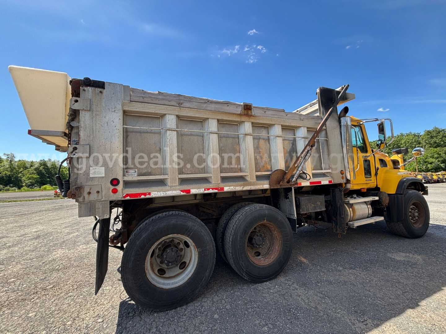 Dump truck side view showing a metal dump bed on a yellow Mack CV713, dual rear axles, with rear wheel on left and tailgate in view.