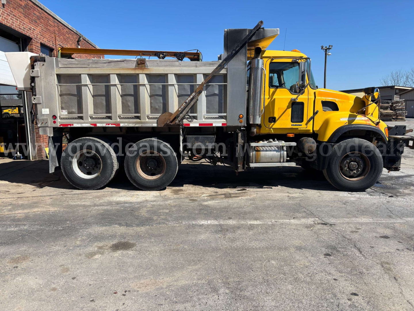 Mack CV713 dump truck side view showing gray dump bed, yellow cab, three rear axles, and hitch mechanism.