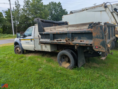 Ford F-550 Super Duty dump truck in in-service condition with rusted dump bed, side cab visible, on grass beside road.