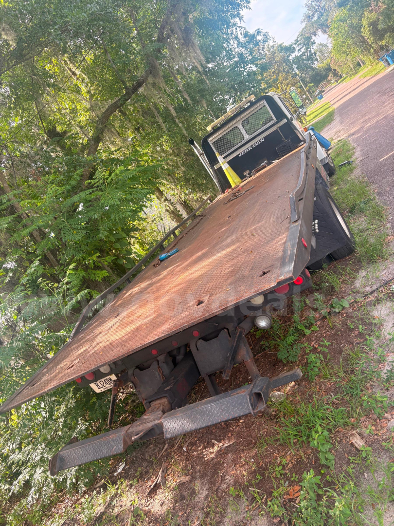 Flatbed tow truck bed and rear cab of a 2003 Freightliner FL80, rusted steel flatbed, visible hitch and bumper, on a grassy roadside with trees.