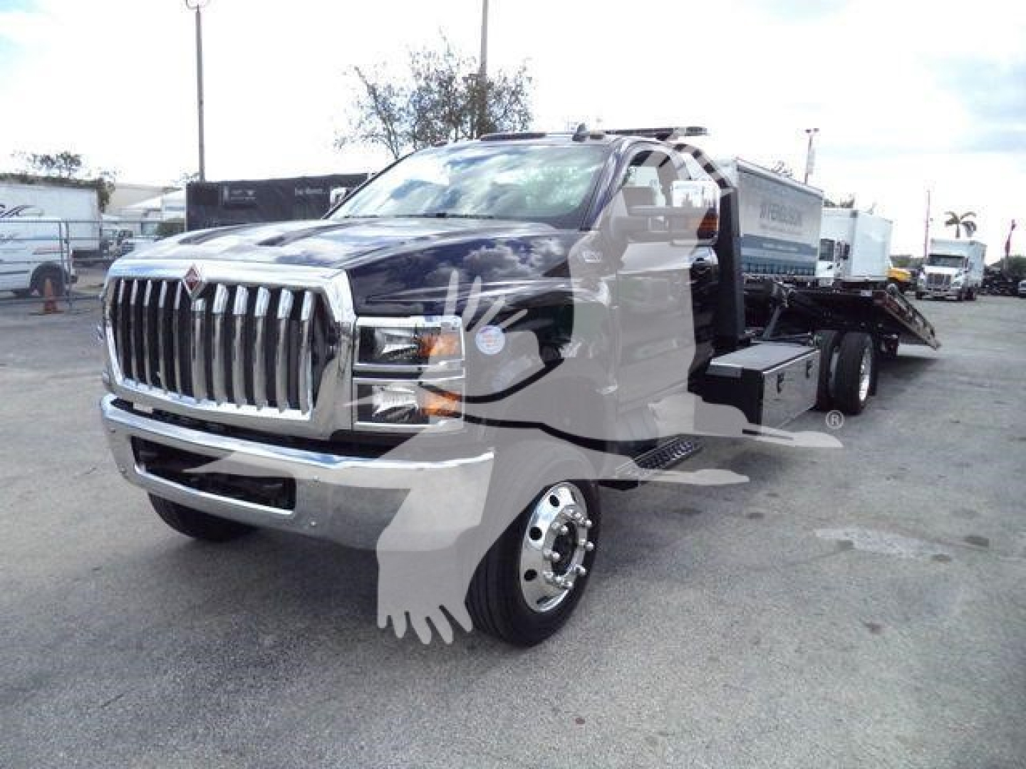 Truck with a chrome grille and flatbed equipment in a dealership lot. Visible text on side panels and grille area; no legible model details beyond general International-style cab and rollback equipment.