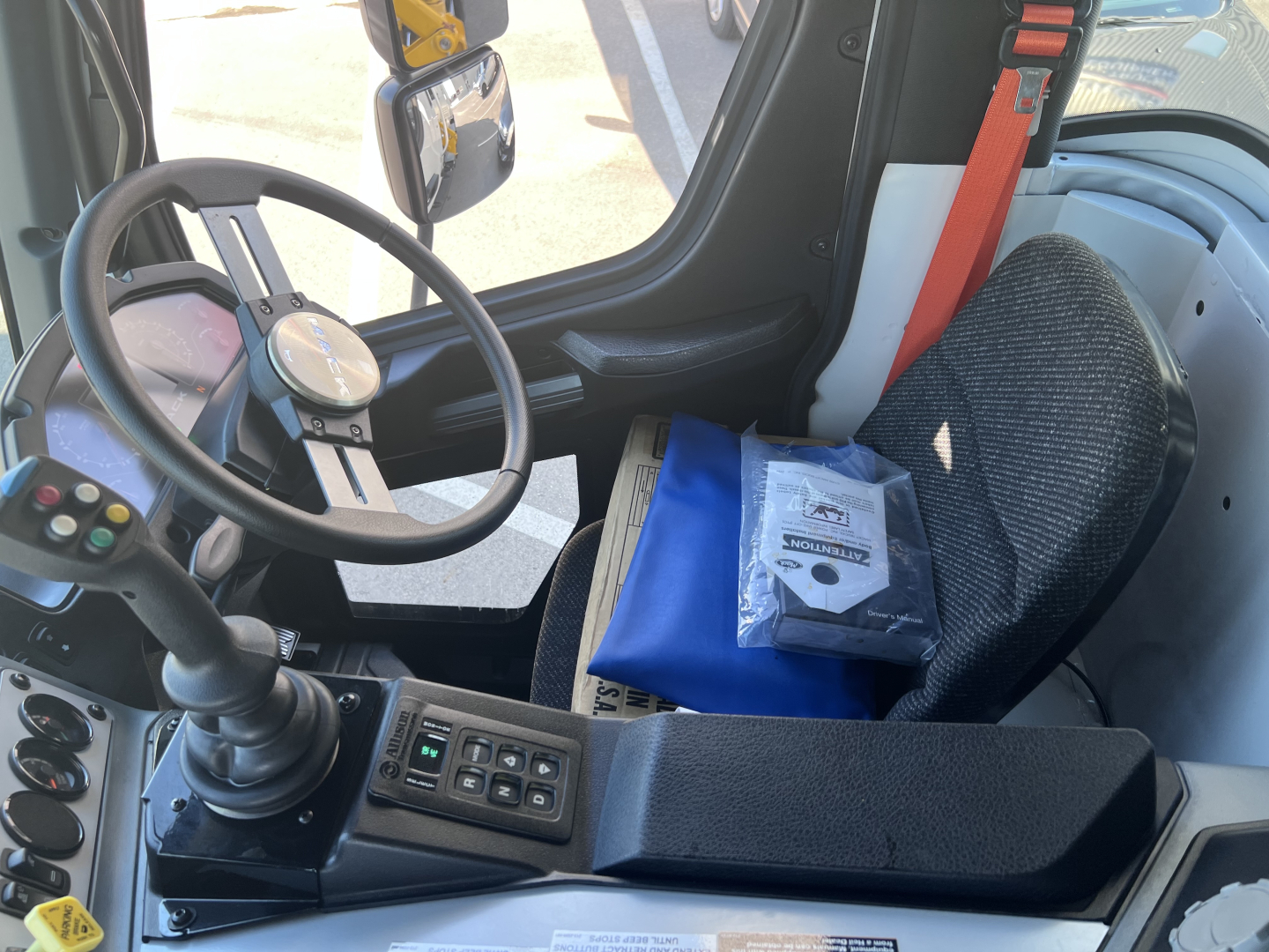 Interior of a cab showing steering wheel with gear shift console and driver seat; control panel with buttons; blue binder and manuals on the armrest.
