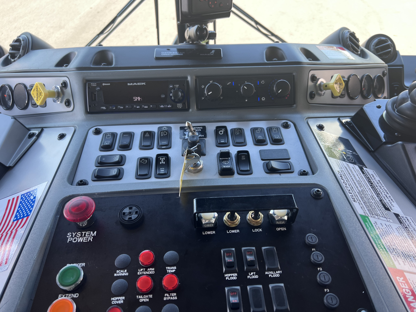 Mack LR64 truck cab dashboard showing numerous switches, gauges, and controls; red and green push buttons; key ignition; steering area; warning stickers visible.
