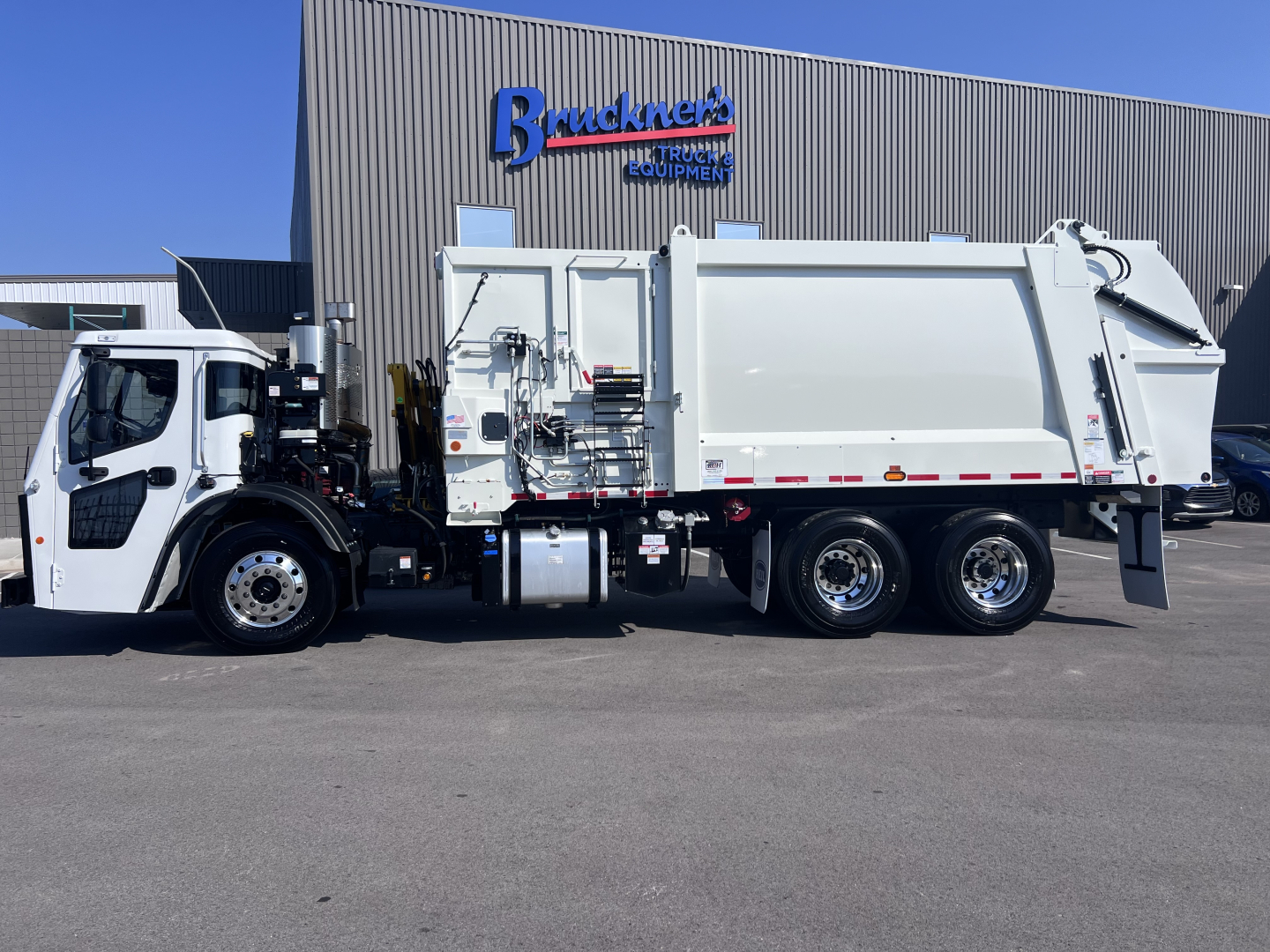 Garbage truck in serviceable condition parked in front of a Mack equipment dealership building with a white cab and body, showing side view clarity.