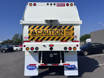 Rear view of a white 2026 Mack LR64 dump truck showing yellow caution sign, taillights, and HEIL branding on mud flaps.