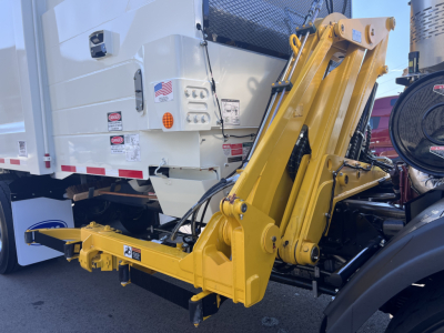 Mack LR64 truck with yellow hydraulic lifting arm attached to rear, side view showing white cab and black fender, no visible text on components.